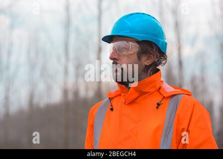 Ritratto di tecnico forestale sicuro in bosco. Uomo professionista adulto che indossa una giacca arancione e un elmetto blu hi-viz blaze. Foto Stock