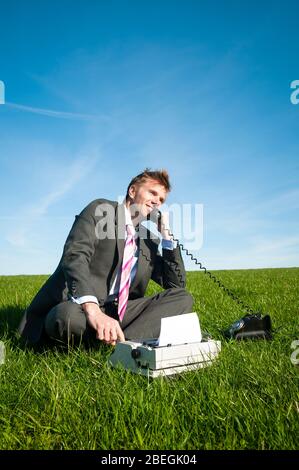 Uomo d'affari sorridente seduto all'aperto con una macchina da scrivere vecchio stile che parla su un telefono a linea rotante Foto Stock