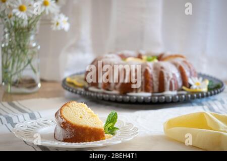 Torta al limone con fiocco di ricotta e glassa, esposta su un piatto decorativo nero con fiori Daisy in un vaso sullo sfondo. Foto Stock