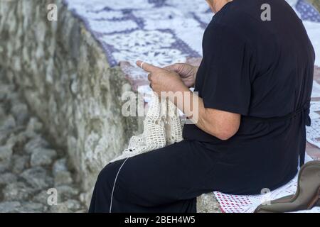 Lavorazione dell'ago, accovacciamento. Una vecchia donna che indossa un abito nero rende tovaglioli di pizzo decorativo con un crochet. Foto Stock
