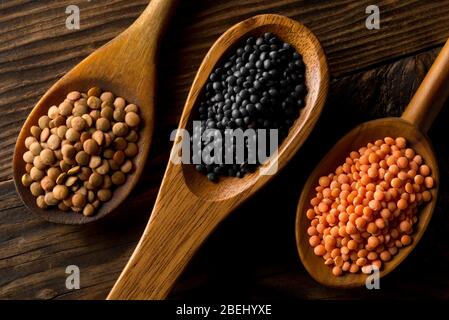 Diverse lenticchie assortite si mescolano con lenticchie beleuga rosse, marroni e nere in scoop di legno su tavola marrone sfondo vista dall'alto piatto posato dall'alto Foto Stock