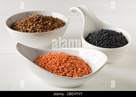 Le lenticchie assortite si mescolano con lenticchie di beluga rosse, marroni e nere in ciotole bianche su sfondo bianco tavolo di legno - fuoco selettivo Foto Stock
