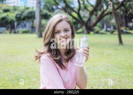 Sorridente giovane donna caucasica ragazza che tiene acqua potabile da una bottiglia di plastica in parco verde Foto Stock