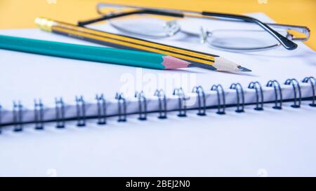 Primo piano di due matite e occhiali su un notebook era aperta pagina vuota, desktop giallo con messa a fuoco morbida. Foto Stock