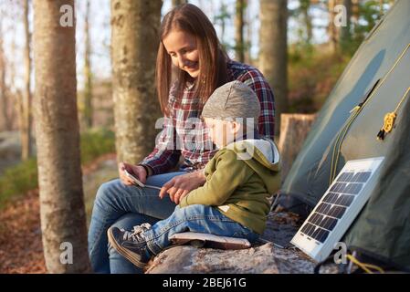 Madre con figlio in vacanza in campeggio guardando un telefono cellulare mentre si gode l'aria fresca. La famiglia siede su un ceppo vicino alla tenda e pannello solare turistico nella foresta in una giornata di sole. Foto Stock