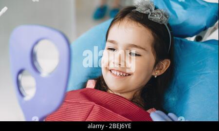 Ragazza piccola felice sorridente nello specchio dopo un intervento chirurgico al dentista Foto Stock