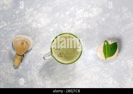 Tazza di vetro con tè giapponese verde matcha, whisky di bambù per montare e foglie su pietre disposte in fila su sfondo grigio testurizzato. Bevanda naturale con Foto Stock