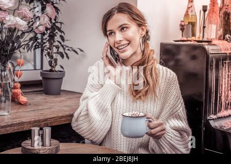 Una donna sorridente felice che parla al telefono bevendo cacao in un accogliente bar Foto Stock