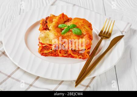 Cannelloni di carne cotti con salsa di pomodoro e mozzarella fusa sopra su un piatto bianco su sfondo di legno bianco, vista orizzontale, primo piano Foto Stock