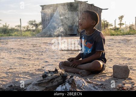 Un giovane ragazzo Ju/hoansi siede accanto al fuoco all'alba nel villaggio di Deng//e a Nyae Nyae, Namibia Foto Stock