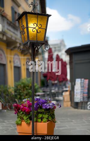 Lampada da strada gialla a fuoco. Bellissimi fiori in vaso. Antichi edifici europei e caffè su una piccola strada italiana come sfondo, sfondo a Pisa Foto Stock