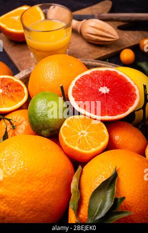 sullo sfondo un bicchiere con succo d'arancia appena spremuto, una spremiagrumi in legno e in primo piano vari tipi di agrumi interi e tagliati a fette Foto Stock