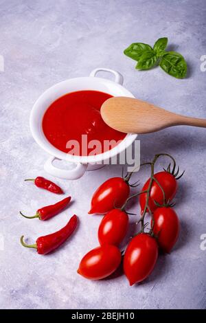 su sfondo grigio rustico, una ciotola bianca in ceramica con salsa di pomodoro, foglie di basilico, pomodori a grappolo e qualche peperone rosso piccante Foto Stock