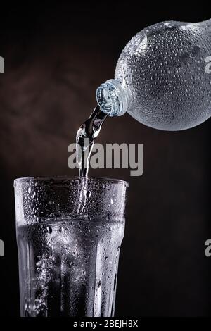 acqua fresca versata da una bottiglia di plastica in un bicchiere di vetro su sfondo scuro Foto Stock