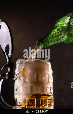 birra fresca e schiumosa versata dalla bottiglia nella tazza di vetro su sfondo scuro Foto Stock