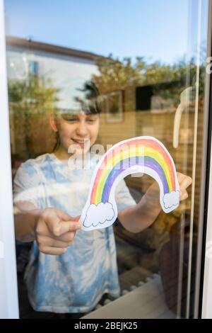 Ragazza che mette l'immagine dell'arcobaleno nella finestra a casa durante la Pandemia di Coronavirus per intrattenere i bambini Foto Stock