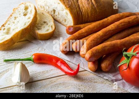 Mazzo di salsicce affumicate sottili, pane affettato, peperoncino piccante, spicchio d'aglio su un tavolo di legno bianco. Salsiccia e verdure fresche come fonte di proteina. Foto Stock