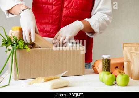 Offrite volontariamente la maschera medica protettiva e i guanti che mettono il cibo nella scatola da donazione. Uomo impiegato in gilet rosso con maschera facciale e guanti di imballaggio Foto Stock