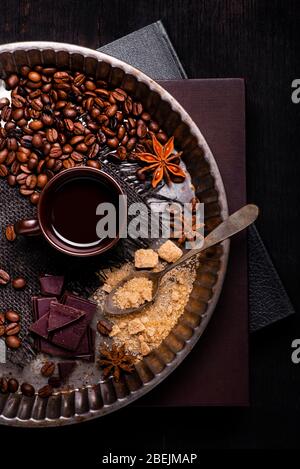 visto dall'alto. sdraiato su alcuni libri, un vassoio con una tazza di caffè nero, un cucchiaio con zucchero di canna, alcuni pezzi di cioccolato fondente e caffè tostato essere Foto Stock
