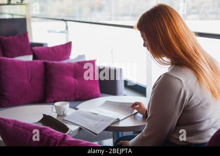 Giovane donna legge una rivista in un bar. Lettura in un luogo pubblico, in una caffetteria, solitudine nel concetto della città Foto Stock