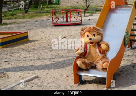 Grande orsacchiotto in un parco giochi deserto. Un orso sorridente si siede su uno scivolo per bambini. Giornata di sole primaverile nei parchi giochi senza persone. Autoisolamento Foto Stock