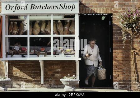 Una donna matura che porta un cestino della spesa lasciando un negozio di macellai locale tradizionale a Orford. Woodbridge, Suffolk, Inghilterra. REGNO UNITO Foto Stock