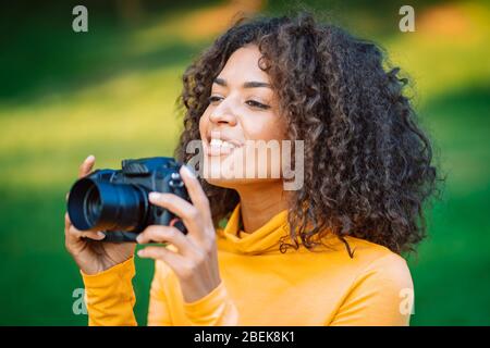 Giovane donna africana graziosa in giallo scatta le immagini con la macchina fotografica di DSLR su sfondo verde. Ragazza sorridente divertirsi come fotografo. Foto Stock