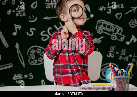 Carino ragazzo intelligente è seduto a una scrivania con lente di ingrandimento in mano. Bambino sta leggendo un libro con una lavagna su uno sfondo. Pronto per la scuola. Indietro Foto Stock