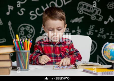 Carino ragazzo intelligente è seduto a una scrivania con lente di ingrandimento in mano. Bambino sta leggendo un libro con una lavagna su uno sfondo. Pronto per la scuola. Indietro Foto Stock