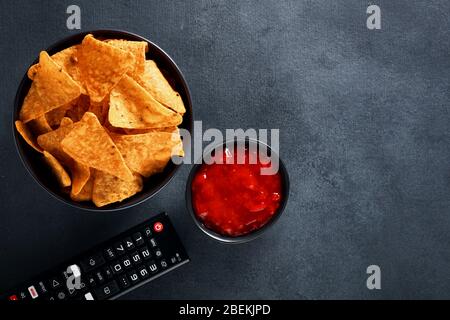 Nachos messicani in una ciotola nera con salsa di salsa calda e telecomando tv su sfondo nero. Guardare la tv e mangiare spuntini. Vista dall'alto con spazio per le copie. Foto Stock