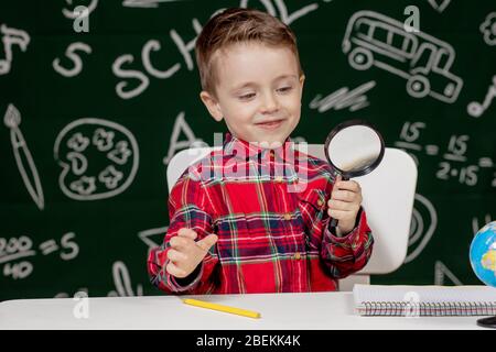 Carino ragazzo intelligente è seduto a una scrivania con lente di ingrandimento in mano. Bambino sta leggendo un libro con una lavagna su uno sfondo. Pronto per la scuola. Indietro Foto Stock