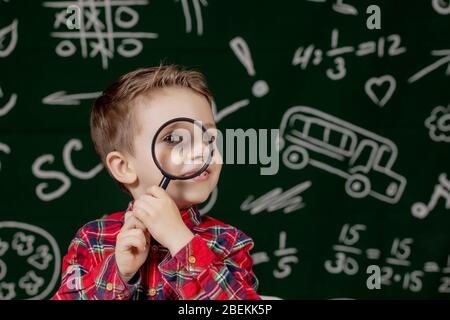 Carino ragazzo intelligente è seduto a una scrivania con lente di ingrandimento in mano. Bambino sta leggendo un libro con una lavagna su uno sfondo. Pronto per la scuola. Indietro Foto Stock