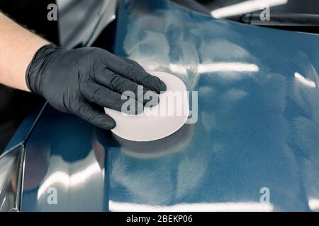 Immagine tagliata ravvicinata della mano maschile nel guanto nero protettivo, che trattiene la spugna bianca con cera carnauba solida e la cappa di lucidatura di auto blu di lusso a. Foto Stock