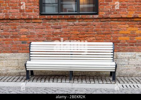 Panca in legno bianco, solitario e vuoto, con parete in mattoni marroni e finestra nera sullo sfondo. Foto Stock