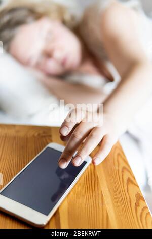 Giovane donna dell'Europa orientale sdraiata a letto e che spegne la sveglia sul suo telefono cellulare al mattino. Foto Stock