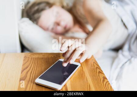 Giovane donna dell'Europa orientale sdraiata a letto e che spegne la sveglia sul suo telefono cellulare al mattino. Foto Stock