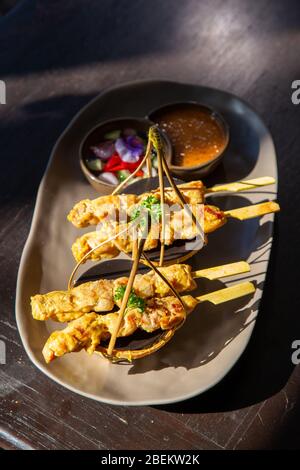 Spiedini di pollo al satay o al kebab su un piatto di ceramica nera con contorni di cetrioli, cipolle rosse, peperoni e salsa tailandese. Foto Stock