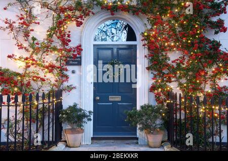 Porta anteriore di una casa incorniciata da una pianta di Pyracantha rampicante decorata con grappoli di bacche rosse e luci di Natale. Poundbury, Dorchester, UK, GB. Foto Stock