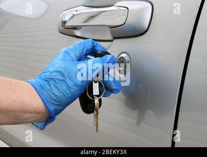 Una vista ravvicinata di una mano in un guanto blu in lattice che sblocca una porta d'auto argentata con chiavi durante la pandemia del coronavirus. Foto Stock