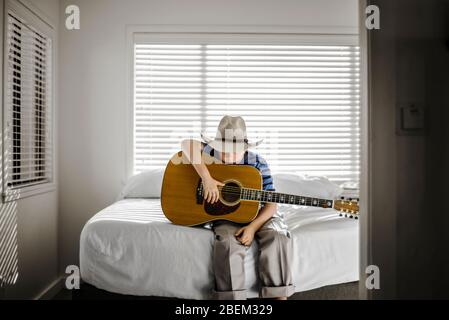 Ragazzo che indossa un cappello da cowboy seduto sul suo letto con una chitarra acustica Foto Stock