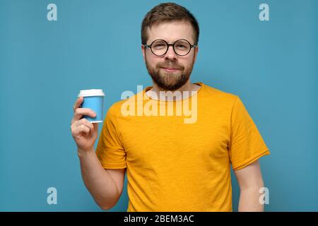 Uomo tranquillo e contented con gli occhiali rotondi e una t-shirt gialla sta tenendo una tazza di carta monouso in mano, su uno sfondo blu. Foto Stock