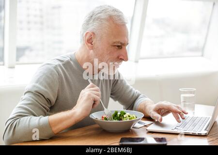 Immagine di un uomo d'affari maturo serio e maturo, con i capelli grigi, che siede al bar, che serve una cena con un computer portatile. Foto Stock