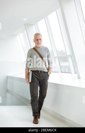 Immagine di un uomo d'affari anziano, barbuto e grigio, che tiene un computer portatile in casa. Foto Stock