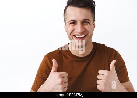 Ritratto ravvicinato di felice carismatico giovane con barba rasata, mostrando i pollici in approvazione o come, sostenere qualcosa di buono, sorridente compiaciuto Foto Stock