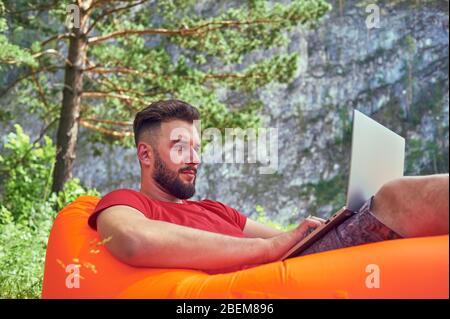 L'uomo bearded sta lavorando e sorridendo con il laptop al parco sotto l'albero. Il freelancer felice è seduto e usando l'app o il sito web sull'erba. Lavoro Foto Stock