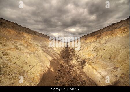 Fossa di drenaggio di campo scavata di fresco che mostra stratificazione di suolo sotto un cielo con nuvole scure Foto Stock