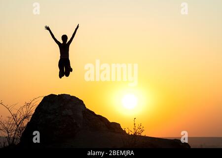 Silhouette di donna escursionista che salta da solo su roccia vuota al tramonto in montagna. Turista femminile che alza le mani in piedi sulla scogliera nella natura serale. Foto Stock