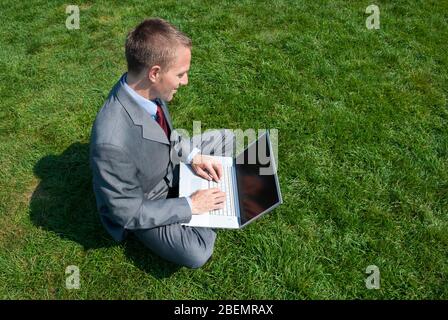 Vista a spalla di un uomo d'affari seduto all'aperto in una zona soleggiata di verde che batte sul suo computer portatile Foto Stock