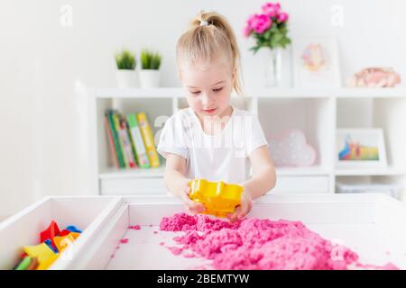 Bambina bionda che gioca con sabbia cinetica rosa su un tavolo bianco in una stanza luminosa. Sviluppo sensoriale. Lezioni in un asilo Foto Stock