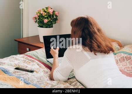 Donna che paga con carta per lo shopping online da casa su un computer portatile, sdraiato sul letto. Autoisolamento. Foto Stock